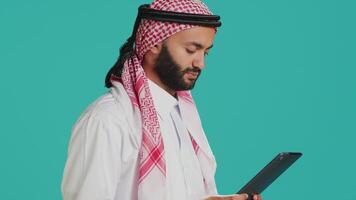 Middle eastern adult browsing device in studio, navigating websites online on digital gadget. Muslim person in traditional clothes using tablet to check internet pages, texting on social media. video
