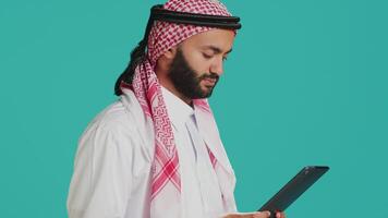 Middle eastern adult browsing device in studio, navigating websites online on digital gadget. Muslim person in traditional clothes using tablet to check internet pages, texting on social media. video