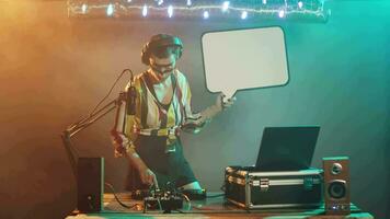 Woman mixer holds speech bubble icon at nightclub, performing techno music mix on stage. Artist using turntables to create mixing show, presenting isolated cardboard placard. Tripod shot. video