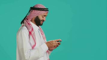 Islamic guy wearing traditional clothes playing online game on smartphone, standing against blue background. Arab gamer enjoying video gaming tournament on mobile phone, entertainment.