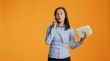 As she chats on retro line, asian woman conducts remote call on her landline phone. Satisfied female model picks up cable office phone and engaging distantly in front of camera. video