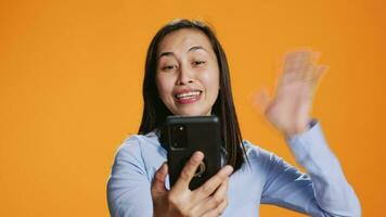 Asian woman answers online videocall in studio, talking to people on internet remote connection. Young adult smiling and waving at phone screen, online videoconference meeting over background. video