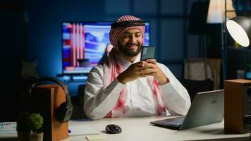 Arabic man at home in living room typing messages on smartphone. Muslim person holding mobile phone, enjoying relaxing leisure time talking with friends over online messaging app photo