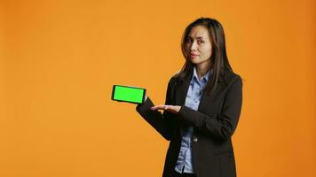 Businesswoman holding phone with greenscreen template, posing against orange background and showing blank copyspace in studio. Asian adult with chromakey screen on smartphone app. video