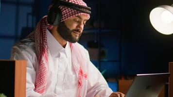 Arabic employee with wireless headphones working from living room office, sending emails. Muslim worker doing internet research using notebook while listening to music to make time pass faster, close up photo