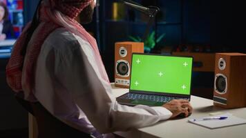 Arabic man scrolling on mockup laptop while working from home. Self employed person writing emails on isolated screen device in apartment office with TV as background noise photo