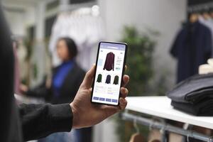 Caucasian customer in a clothes store is holding a smartphone and looking at clothes on a boutique website. Male shopaholic is perusing clothing items in a mall using a cellphone. photo