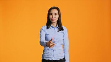 Filipino person showing thumbs up sign on camera, expressing good vibes and positivity over orange background. Young smiling woman giving like in studio, presenting approval gesture. photo