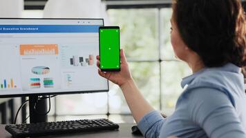 Employee holding mobile phone at desk, looking at isolated greenscreen layout presented on smartphone device. Businesswoman working with modern software technology, chromakey mockup. photo