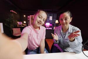 Lively child and her mum do content creation using selfie camera to film vlog channel intro. Daughter and parent greet fanbase, presenting today video topic in pink neon illuminated studio, POV shot photo