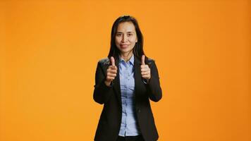 Filipino employee does thumbs up gesture on camera, expressing positivity with like symbol over orange background. Young woman in formal attire presenting approval sign in studio. photo