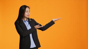 Asian employee points at left or right sides in studio, creating new web commercial over orange background. Young business manager working on marketing advertisement during shoot. photo