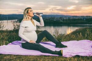 Beautiful pregnant woman enjoys drinking water while spending time outdoor. photo