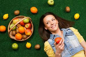 Beautiful young woman is eating apple while lying down on the grass with a lot of fruit around her photo