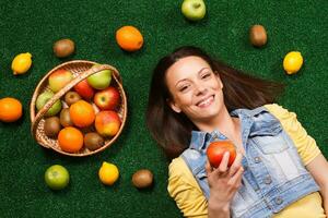 Beautiful young woman is eating apple while lying down on the grass with a lot of fruit around her photo