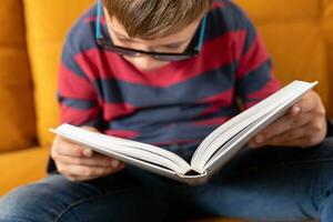 Inquisitive young boy with glasses engrossed in reading a book on the couch photo