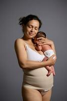 Loving caring multiethnic young woman mother carrying and hugging her newborn baby, isolated over gray studio background photo