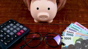 Piggy bank with glasses, calculator, and stacks of money, indonesian currency on wooden background video