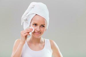 Beautiful young woman cleaning her face with cotton pad on gray background. photo