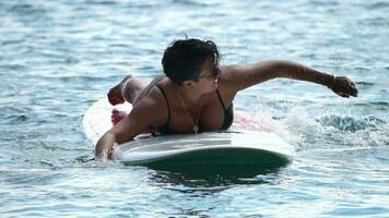 Happy body positive woman smiling and paddling on her surfboard in ocean or sea to beach after surfing on sunny day. Modern individual female hipster outdoor summer sport activity. Slow Motion video