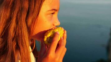 Happy girl eating corn. Summer snacking on the sea. Portrait of young beautiful woman eating grilled corn while sitting by the sea on sunset time. Close up. Slow motion. video