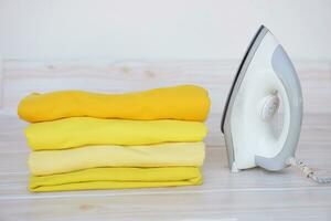 Stack of folded yellow clothes and electric iron on table. Concept, daily chore, household. ironing and folded clothes for neat and clean. Maintenance and keep garments. photo