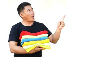 Handsome Asian man hold folded colorful clothes, point finger up with funny face expression. Excited.White background. Concept, daily chore, household. Folded clothes for neat and clean. Keep garment. photo