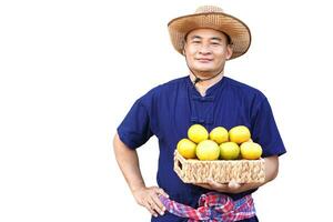 Handsome Asian man farmer wears hat, blue shirt, put hand on waist and holds basket of fruits, isolated on white background. Concept, Agriculture occupation, produce crops to market. photo