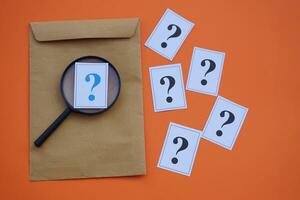 Brown envelope,magnifying glass and paper cards of question marks,orange background. Concept. Teaching aid. Education materials for doing activity or playing investigation games about find answers. photo