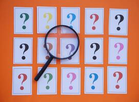 Paper cards of colorful question marks and magnifying glass on orange background. Concept. Teaching aid. Education materials for doing activity or playing investigation games about finding answers. photo