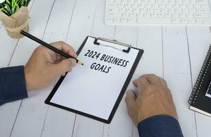 Hands writing a 2024 business goals on a clip board on white wooden desk. photo