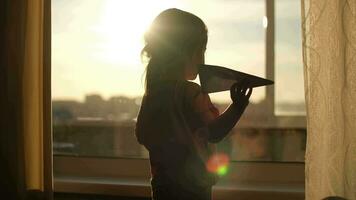 A child plays with a paper plane near the window of the house during sunset.Authentic scene. video