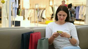 feliz mulher viciado em compras blogueiro usa Móvel telefone enquanto sentado dentro Shopping perto boutique com roupas. video