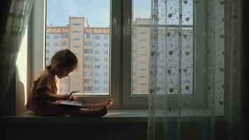 Kind Mädchen Alter 2 Jahre alt Verwendet ein Tablette während Sitzung auf das Fensterbrett beim home.education beim heim. Stadt Leben. sonnig Morgen. video