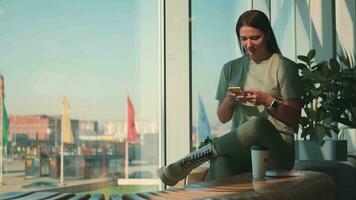 Business woman in casual clothes writes a message using a mobile phone while sitting near a panoramic window on the background of the city. video