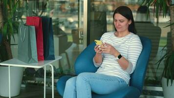 Happy female shopaholic blogger uses mobile phone while sitting in the mall. video