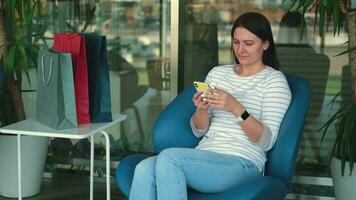 Happy female shopaholic blogger uses mobile phone while sitting in the mall. video