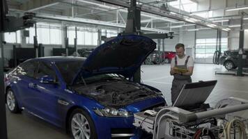 Mature male mechanic making notes and using computer while repairing a car video