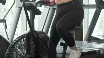 Beautiful curvy woman smiling to the camera doing cardio on air bike at gym video