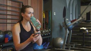 a desportivo menina parece para dentro a telefone e bebidas água durante uma pausa entre exercícios video