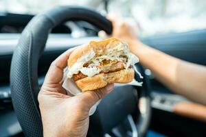 Asian lady holding hamburger to eat in car, dangerous and risk an accident. photo