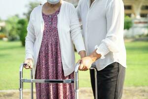 Caregiver help Asian elderly woman patient walk with walker in park, healthy strong medical concept. photo