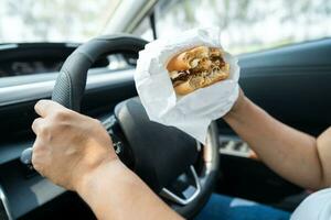 Asian lady holding hamburger to eat in car, dangerous and risk an accident. photo