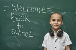 a boy standing in front of a blackboard with the words welcome back to school written on it photo