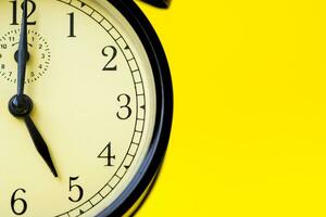 Closeup of a vintage analog black clock on a yellow background displaying 5 o'clock photo