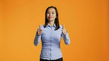 Asian model presenting thumbs up symbol in studio, expressing good vibes and success against orange background. Young smiling person giving like sign on camera, shows approval gesture. video