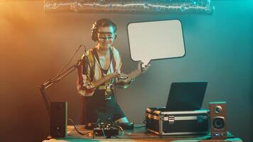 Woman mixer holds speech bubble icon at nightclub, performing techno music mix on stage. Artist using turntables to create mixing show, presenting isolated cardboard placard. Tripod shot. photo