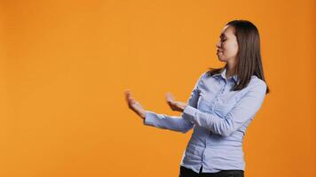 Asian person presenting something to studio sides, creating new marketing advertisement posing over orange background. Young woman pointing left or right for web promotional ad. video