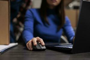 Woman managing parcel delivery on laptop in retail ecommerce warehouse. Logistics manager doing inventory check on laptop while using computer mouse with close up on hand photo