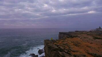 einsam Mann im Gelb Jacke ist Stehen auf das Kante von Cliff und suchen auf das atlantisch Ozean auf wolkig Abend. Peniche. Portugal. Antenne Sicht. Drohne bewegt sich nach vorne video